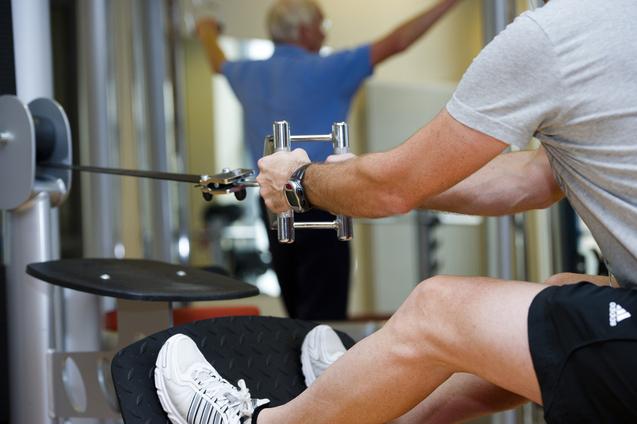 Mann trainiert im Fitnessstudio mit einem Kabelzuggerät, ein anderer Mensch im Hintergrund führt eine Übung aus.