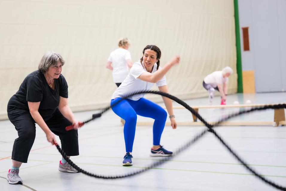 Zwei Frauen trainieren mit einem Battle Rope in einer Sporthalle, mehrere weitere Personen im Hintergrund aktiv.