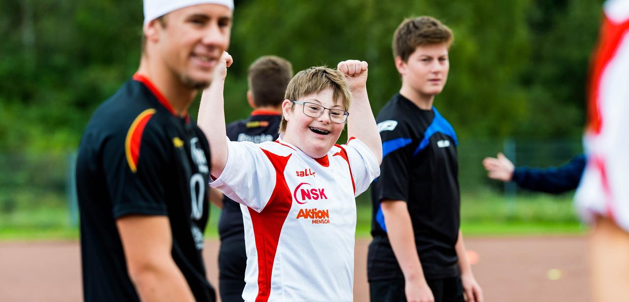 Ein junger Spieler in einem weißen Trikot hebt die Arme jubelnd, umgeben von weiteren Fußballspielern auf dem Platz.