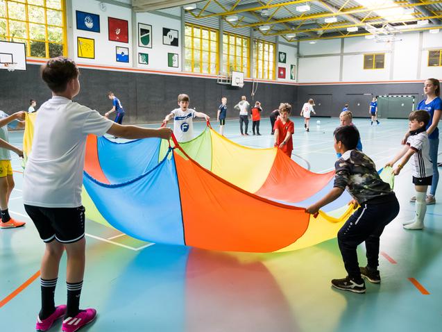 Gruppe von Kindern in einer Sporthalle, die einen bunten Parachute in die Höhe halten und gemeinsam spielen.