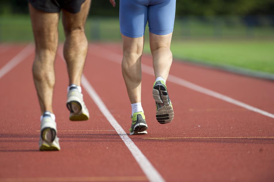 Zwei Läufer, von hinten gesehen, joggen auf einer roten Laufbahn. Der eine trägt kurze Sportshorts, der andere eine blaue Hose.