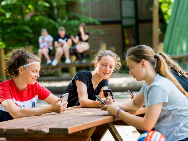 Vier Jugendliche sitzen an einem Holztisch im Freien und schauen auf ihre Smartphones.
