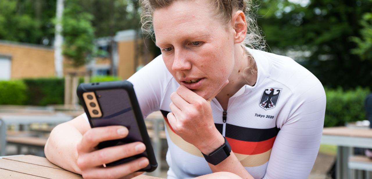 Athletin im weißen Trikot mit schwarzem und rotem Streifen schaut konzentriert auf ihr Smartphone in einer Außenanlage.