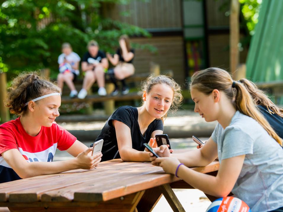 Vier Jugendliche sitzen an einem Holztisch im Freien und schauen auf ihre Smartphones.