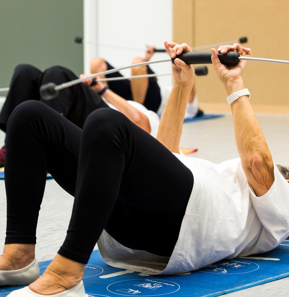 Ältere Personen machen auf Gymnastikmatten Liegestütze mit Widerstandsband in einem Trainingsraum.