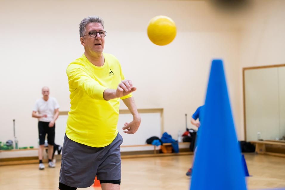 Ein Mann in gelber Sportkleidung wirft einen Ball auf einen blauen Kegel in einem Übungsraum. Hintergrund unscharf.