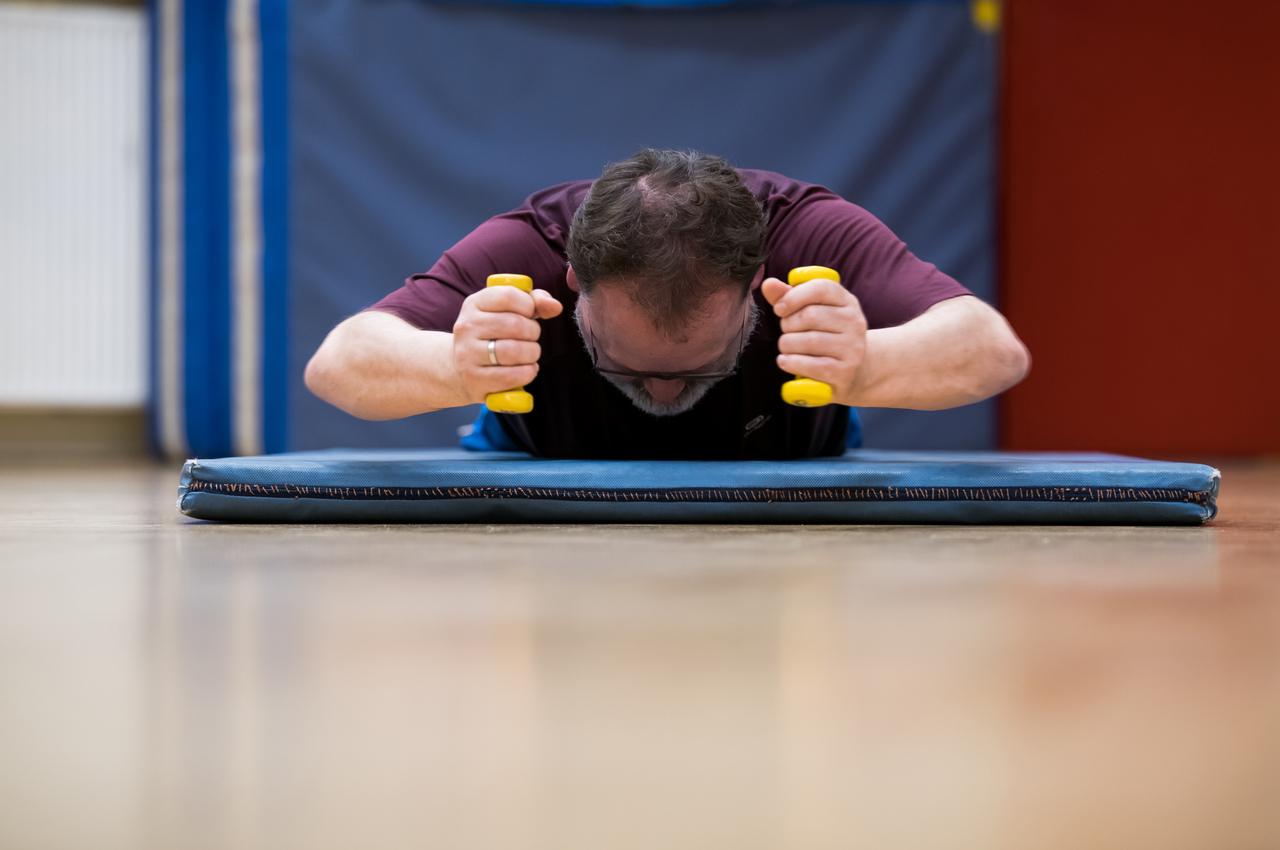 Mann trainiert auf einer Matte mit gelben Hanteln, Kopf nach unten, in einem Sportraum mit blauen Wänden.