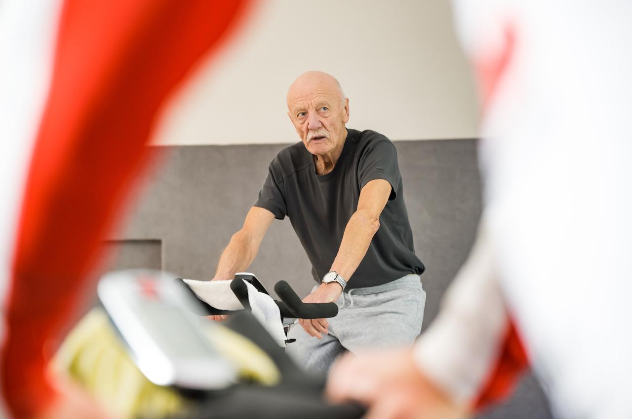 Älterer Mann auf einem Spinning-Bike, konzentriert, mit Hand auf dem Lenker und Handtuch auf dem Sattel.
