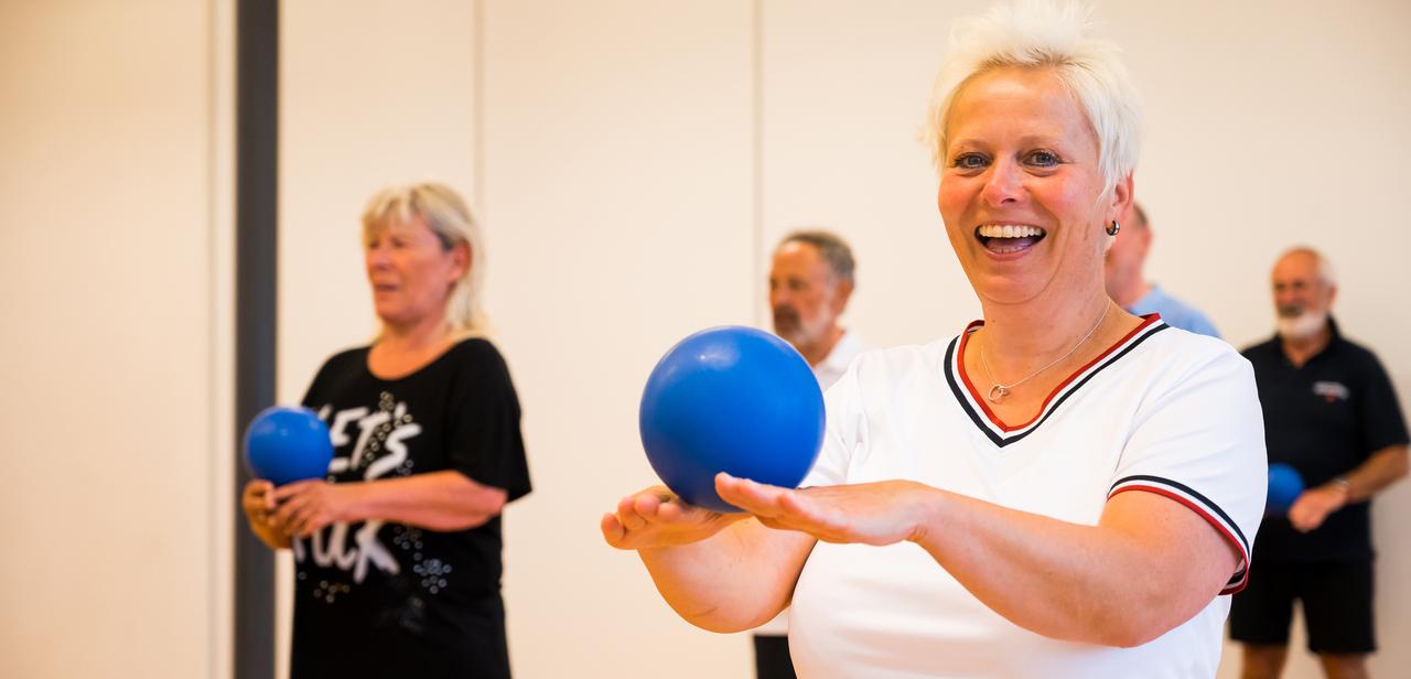 Gruppe älterer Menschen bei einer Übung mit blauen Bällen in einem hellen Raum. Fokus auf lächelnde Frau vorne.
