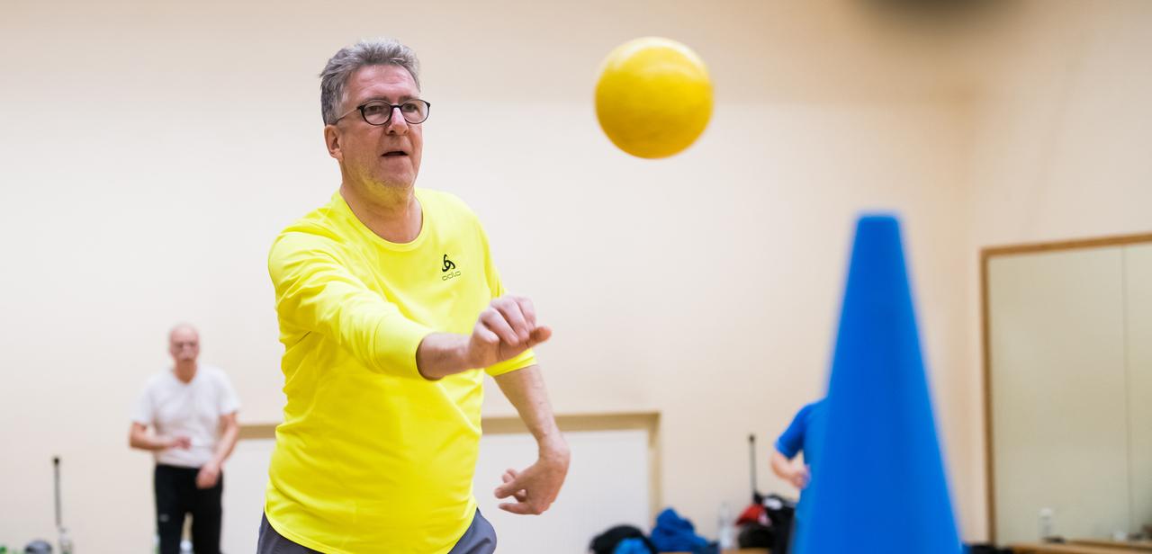Ein Mann in gelber Sportkleidung wirft einen Ball auf einen blauen Kegel in einem Übungsraum. Hintergrund unscharf.