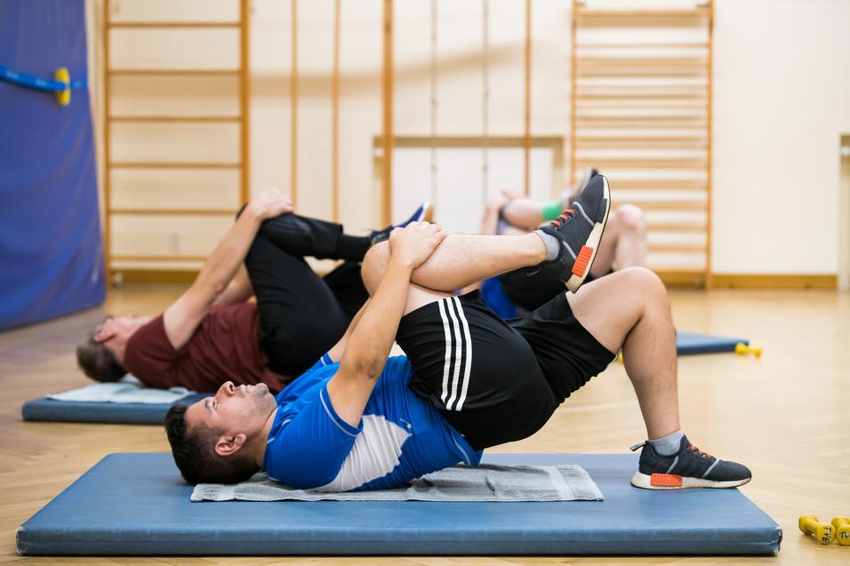 Zwei Männer machen eine Rückenübung auf Matten in einem Trainingsraum, jeder hält ein Bein zu sich hin.