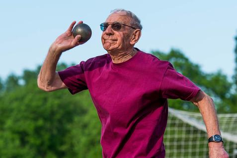 Älterer Mann in kurzer Sportkleidung wirft eine Kugel auf einem Sportplatz mit Fußballtor im Hintergrund.
