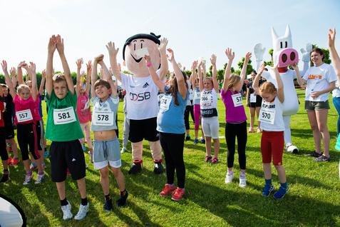 Kinder mit Nummern auf ihren T-Shirts stehen auf einer Wiese und heben die Arme, umgeben von Maskottchen.