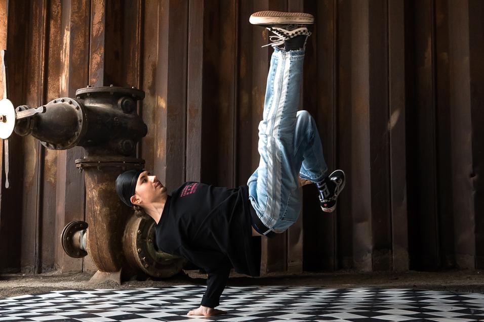 Junge in sportlicher Kleidung führt einen Handstand auf einem schachbrettartigen Boden in einer industriellen Umgebung aus.