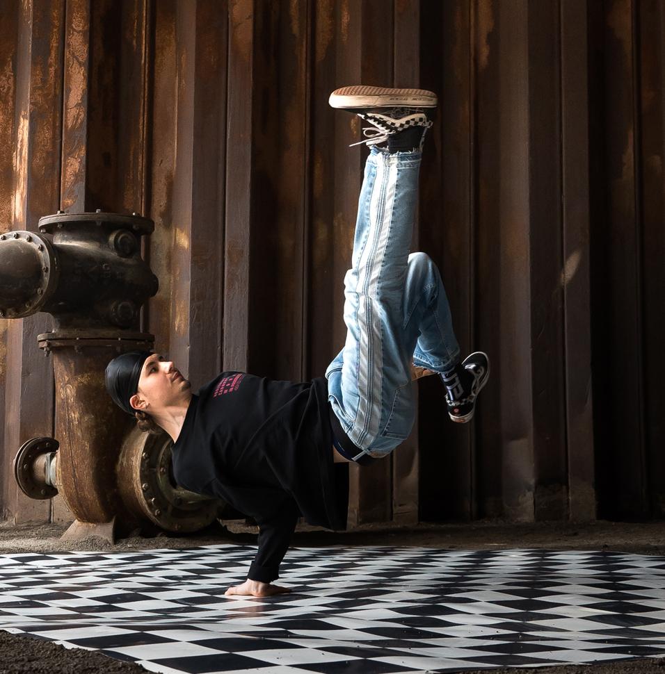 Junge in sportlicher Kleidung führt einen Handstand auf einem schachbrettartigen Boden in einer industriellen Umgebung aus.