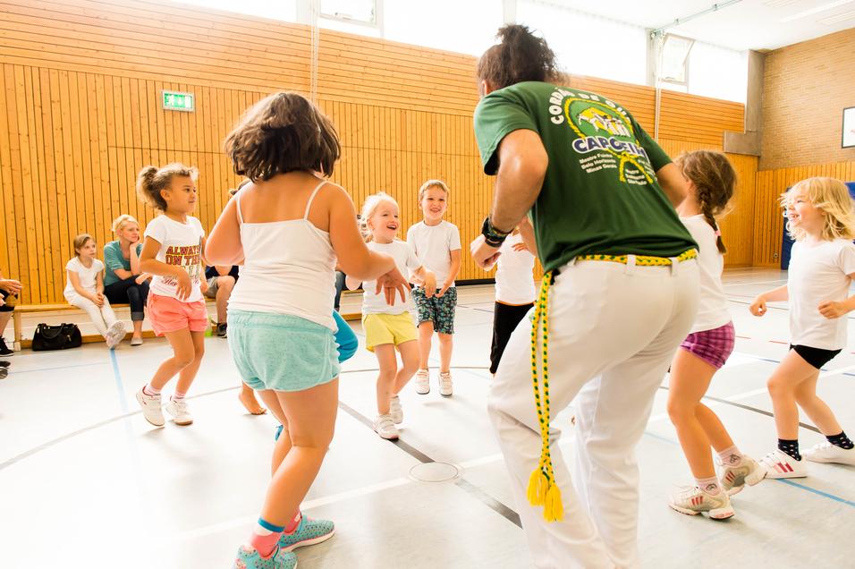 Gruppe von Kindern tanzt fröhlich in einer Turnhalle, geleitet von einem Trainer in grünem T-Shirt.