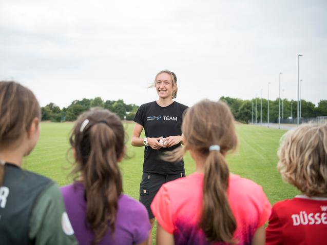 Trainerin spricht mit jungen Sportlerinnen auf einem grünen Fußballfeld unter bewölkten Himmel.