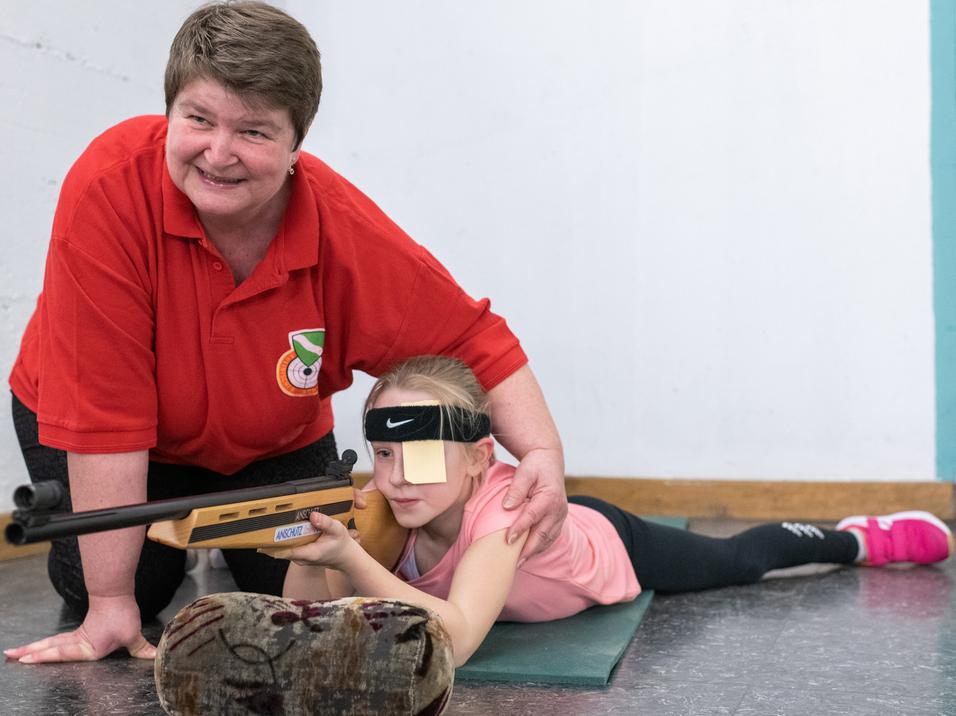 Eine Trainerin unterstützt ein Mädchen beim Schießen, während es auf einem Teppich liegt und zielt.