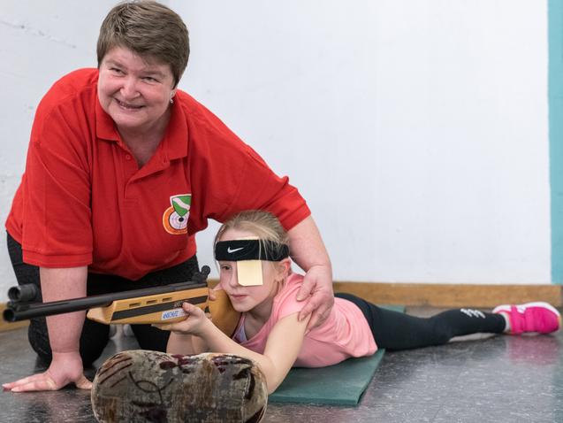 Eine Trainerin unterstützt ein Mädchen beim Schießen, während es auf einem Teppich liegt und zielt.