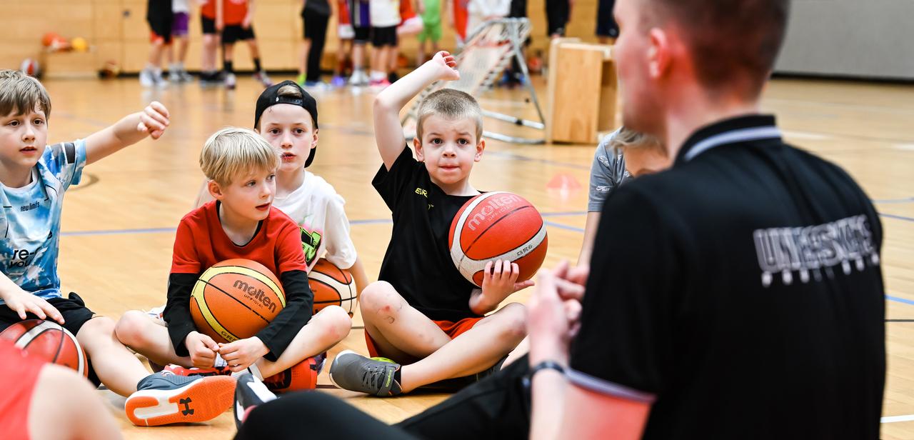 Eine Gruppe von Kindern sitzt auf dem Boden der Sporthalle und hält Basketballs, während ein Trainer ihnen zuschaut.
