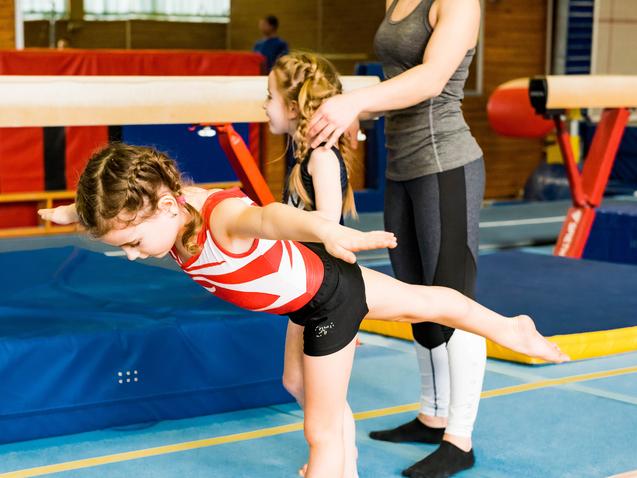 Zwei Mädchen trainieren in einer Turnhalle; eines balanciert auf einem Schwebebalken, während das andere unterstützt.