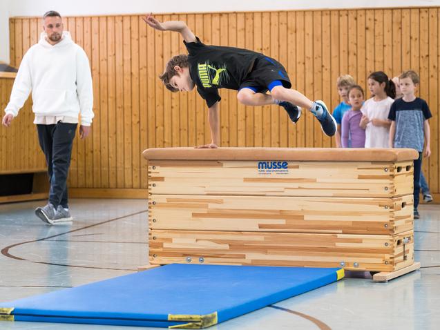 Ein Junge springt über ein Turngerät, während eine Gruppe Kinder und ein Trainer zuschauen. Sporthalle mit Holzverkleidung.