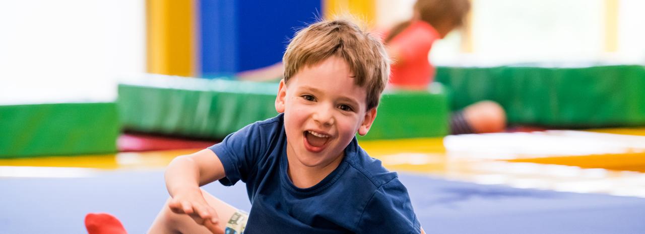 Ein fröhlicher Junge spielt auf einer blauen Matte in einer Freizeitstätte, umgeben von bunten Matten.