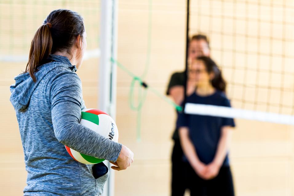 Eine Person mit einem Volleyball steht vor dem Netz, während zwei weitere Personen im Hintergrund warten.